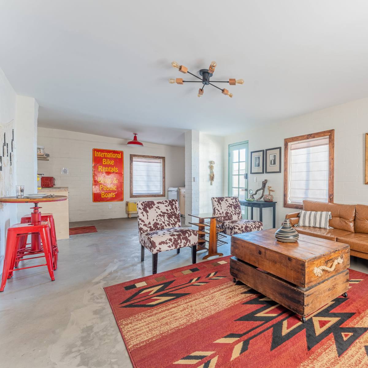 The room features a combination of a leather sofa, patterned chairs, a wooden coffee table, and a colorful area rug, with windows allowing natural light to illuminate the space.