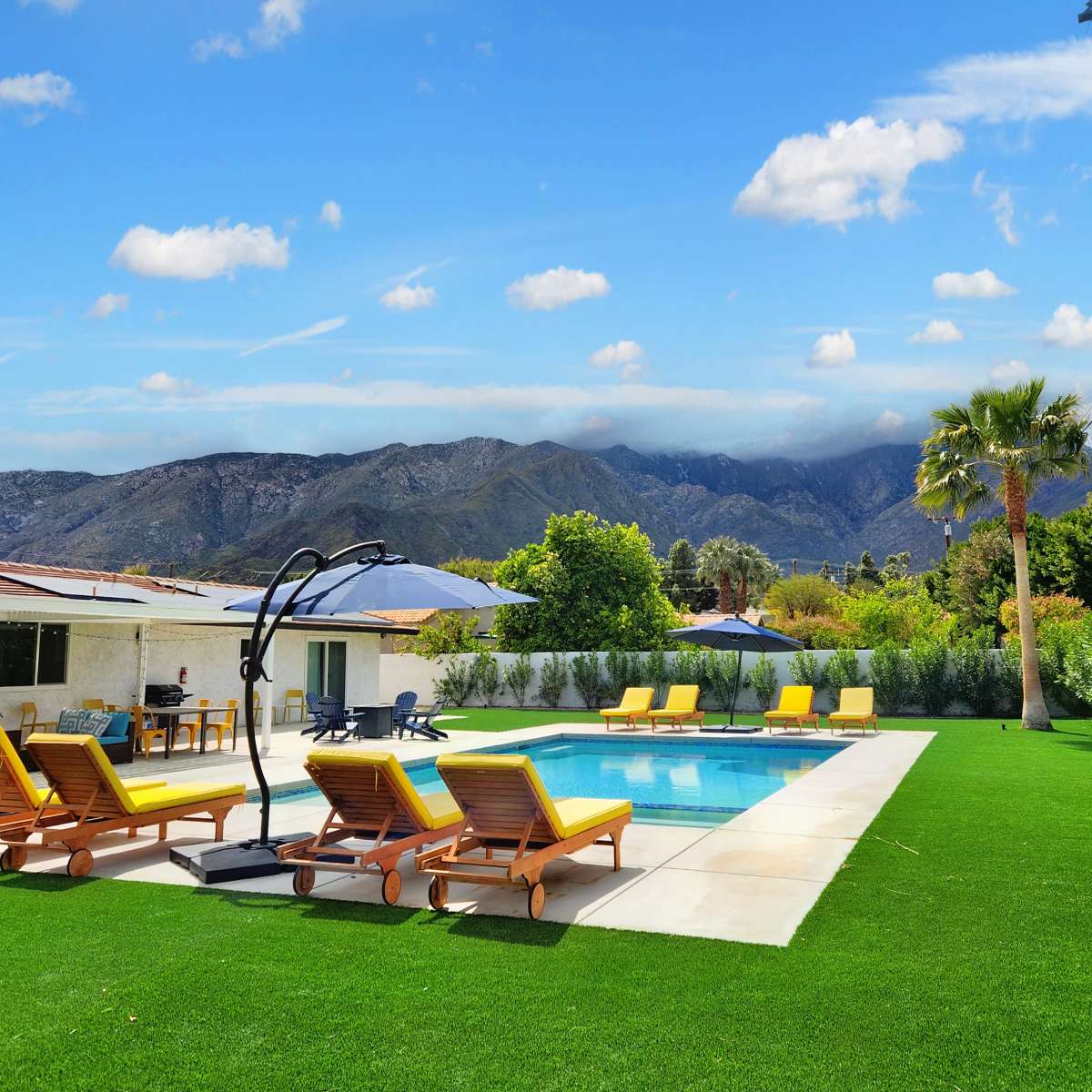 A landscaped backyard features a swimming pool surrounded by yellow lounge chairs, with mountains and palm trees in the background.