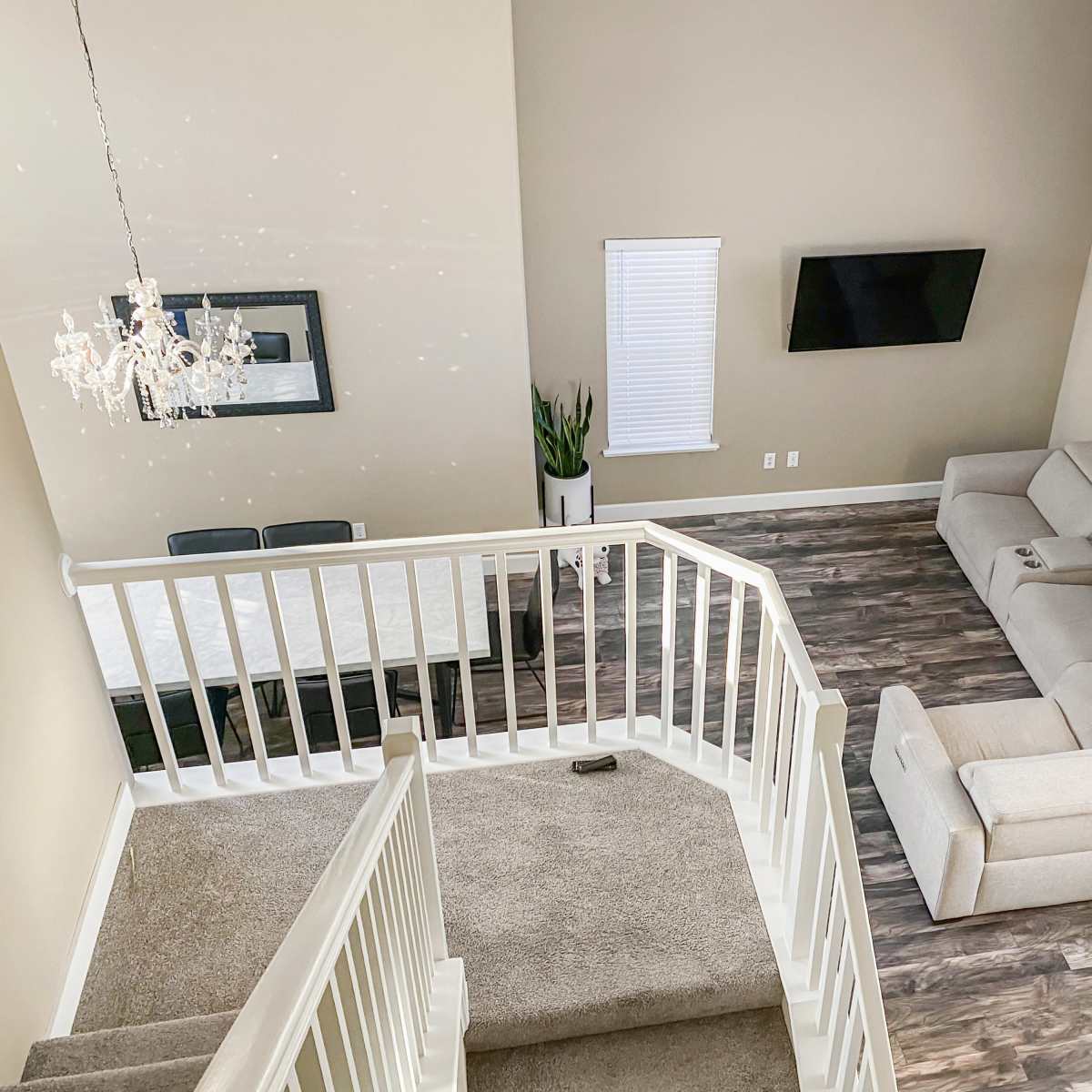 A spacious living room features a light-colored sectional sofa, a flat-screen TV mounted on the wall, and a dining area with a table, all viewed from a balcony overlooking the scene.