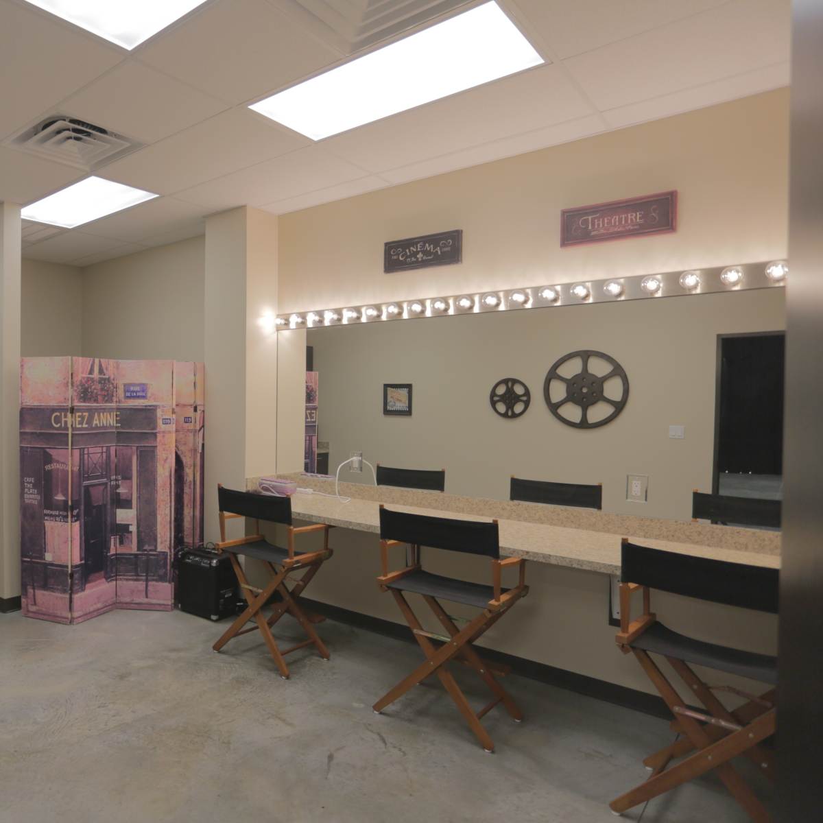 A spacious room with a kitchenette area on one side and a mirrored vanity with illuminated bulbs and director's chairs on the other.