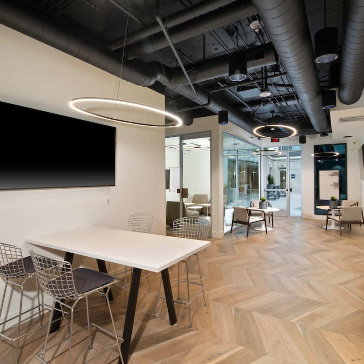 A modern office interior featuring a communal workspace with a table and chairs, sleek lighting, and an open layout with glass-walled conference rooms.