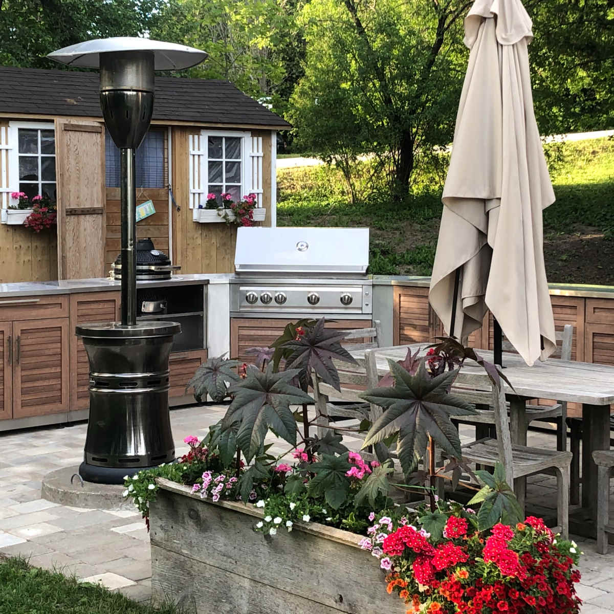 An outdoor kitchen area with a stainless steel grill, wooden cabinets, a patio table with chairs, a large umbrella, and a decorative planter filled with flowers.