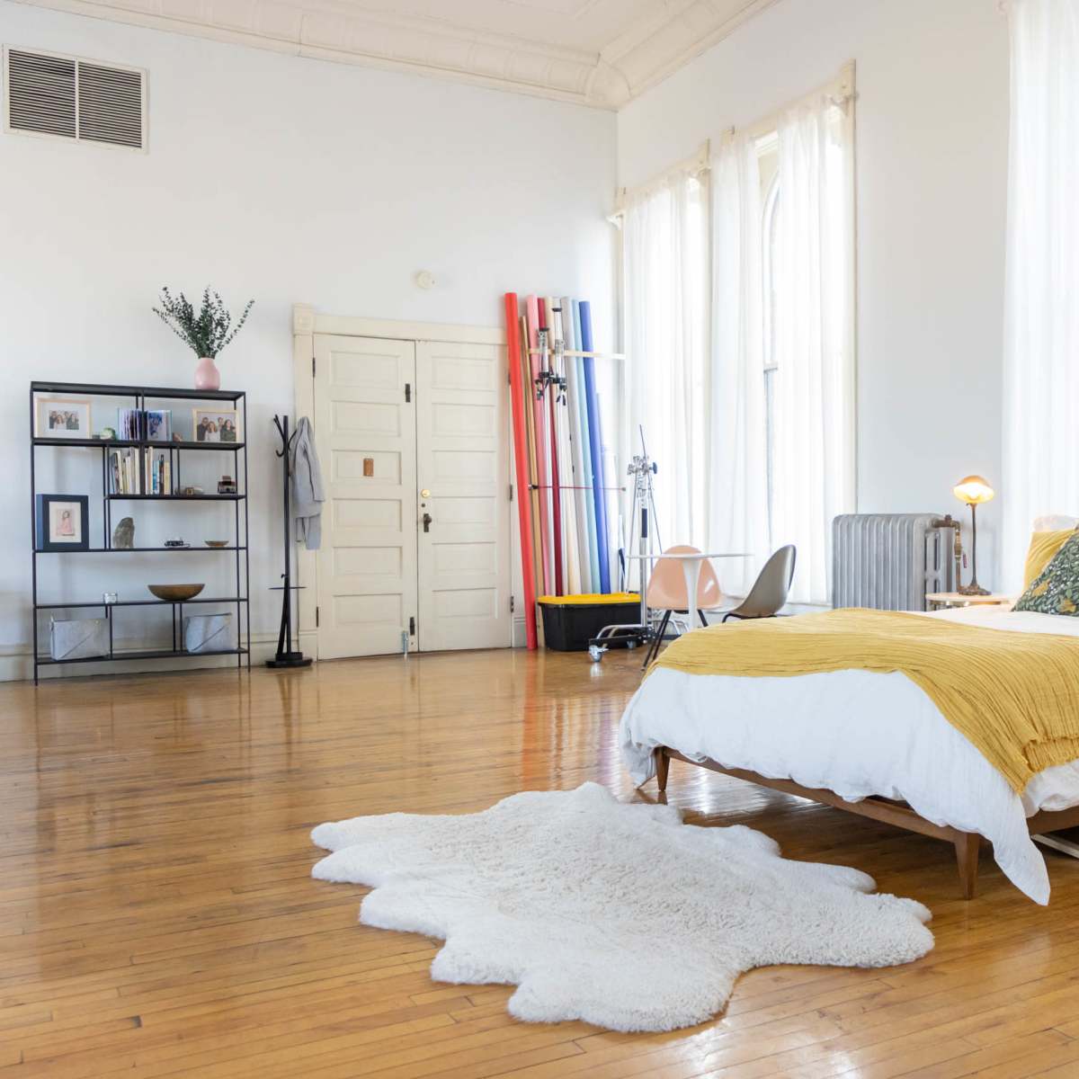 A bright, minimalistic bedroom with a wooden bed, a fluffy white rug on the polished wooden floor, and large windows allowing natural light to fill the space.