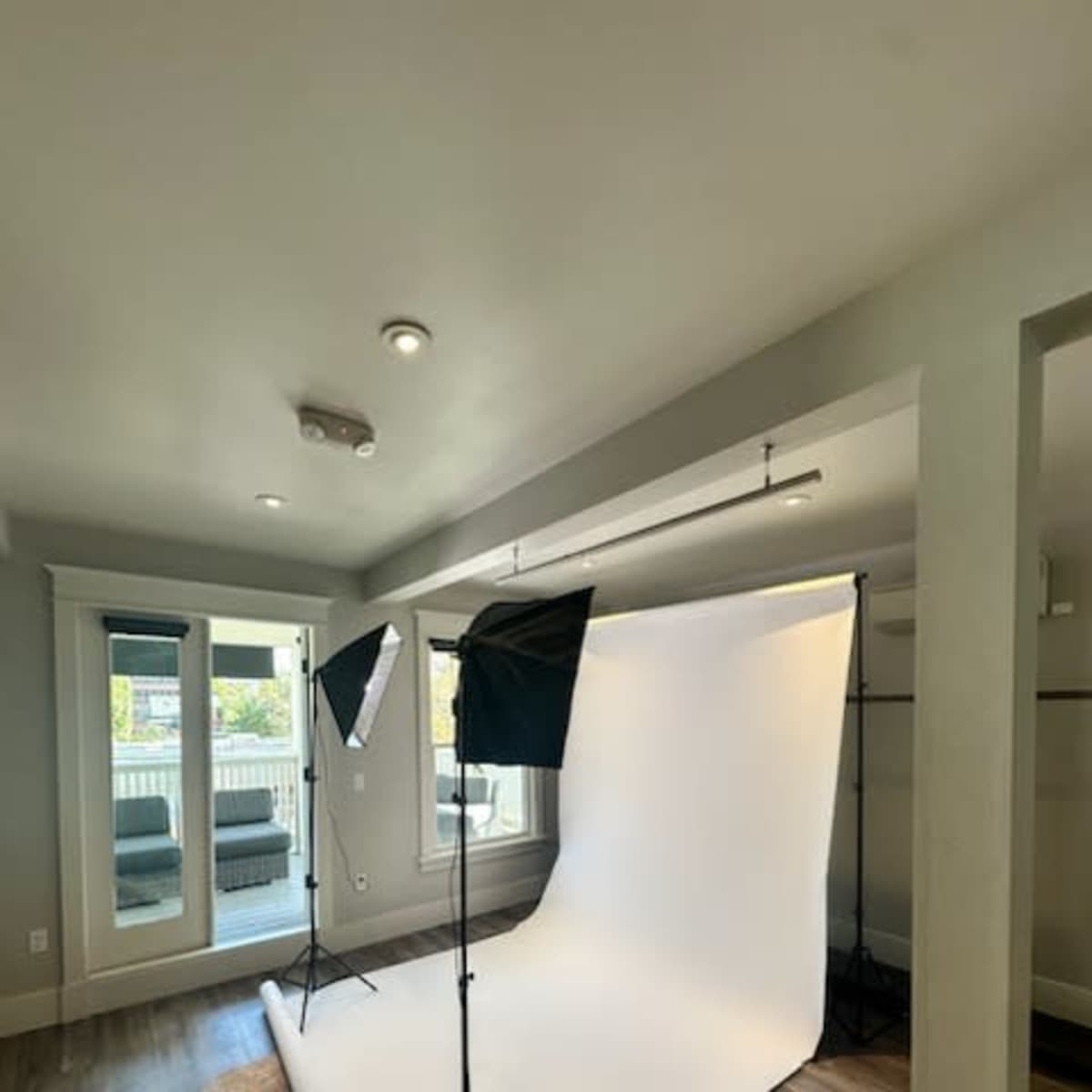 A photography studio setup featuring a white backdrop, two lighting softboxes, and a view of a patio area through sliding glass doors.