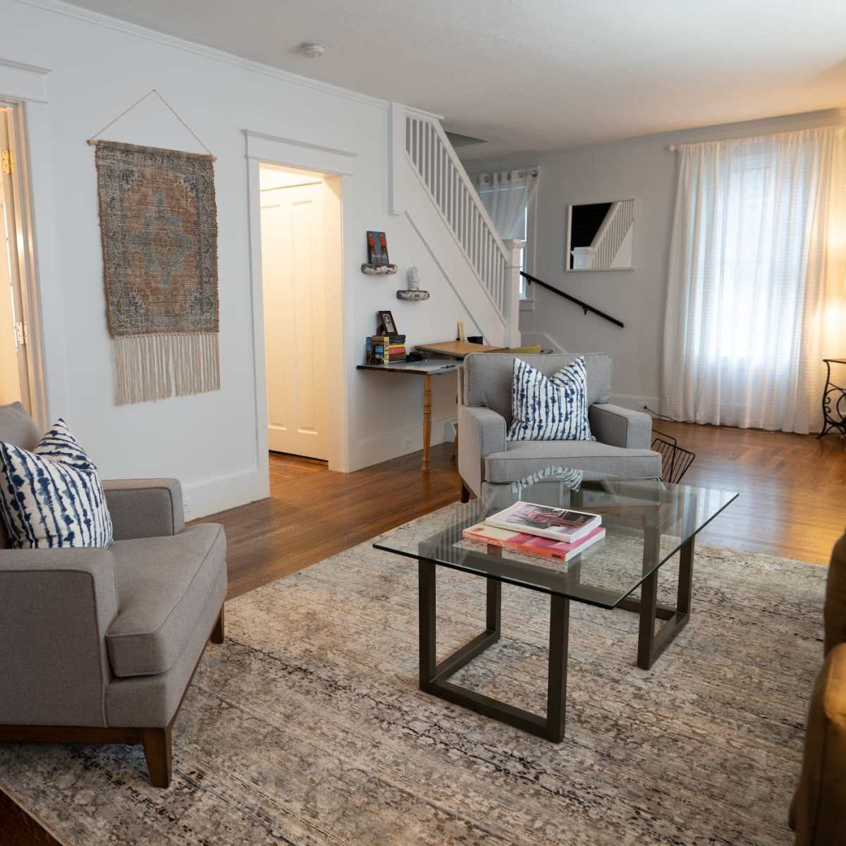 The living room features two gray armchairs facing a glass coffee table on a patterned rug, with an open doorway leading to a dining area and a staircase in the background.