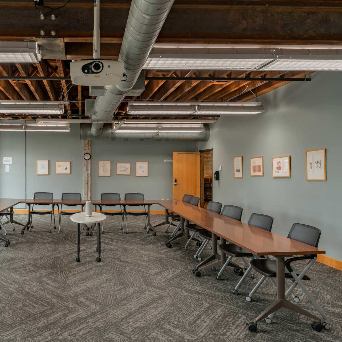 A spacious meeting room features a U-shaped arrangement of tables and chairs with exposed brick walls and overhead lighting.