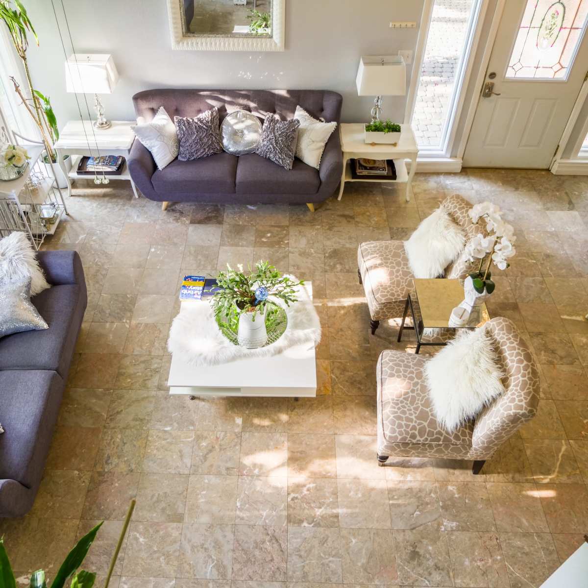 A well-lit living room with two sofas, a coffee table, and decorative plants arranged on a tiled floor.
