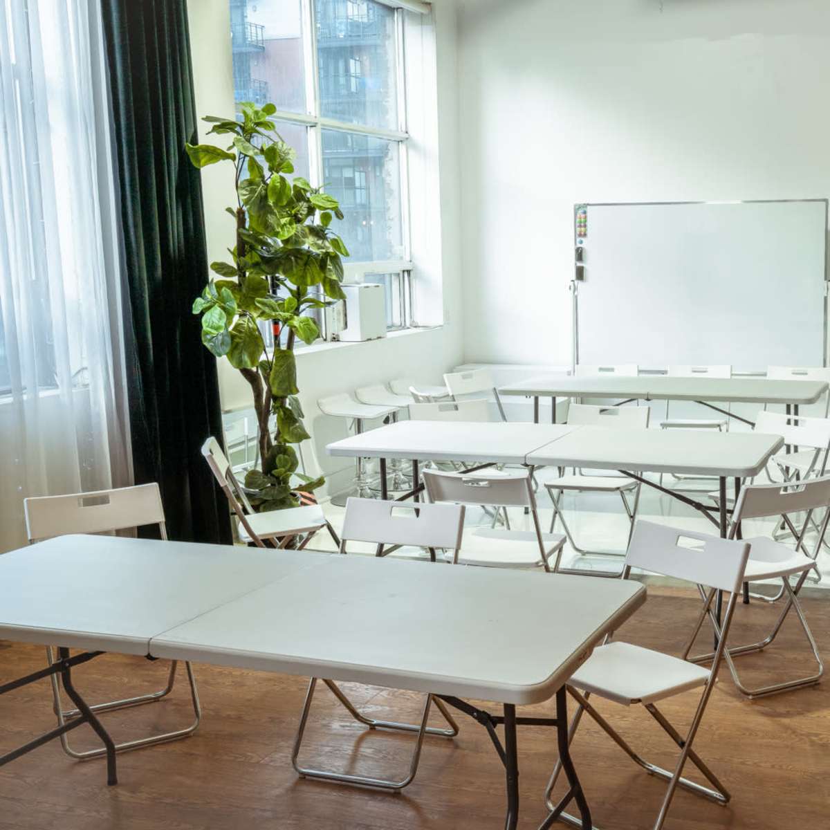 A bright classroom is arranged with several folding tables and chairs, alongside a whiteboard and a television on the wall.