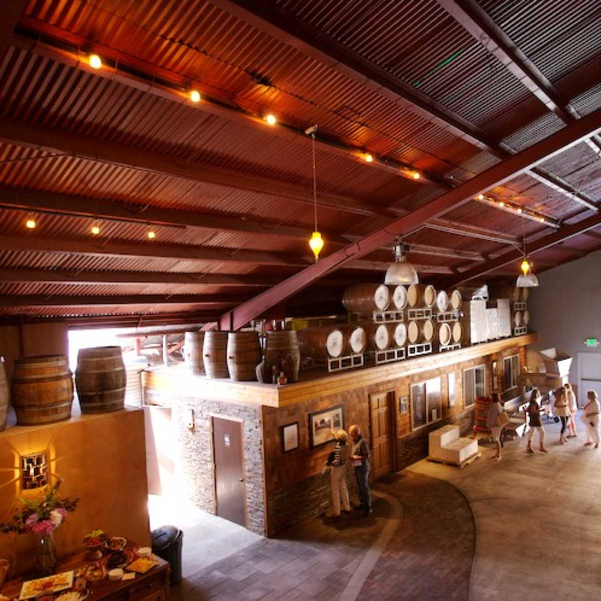A spacious winery interior with high ceilings, wooden barrels, and several people engaged in conversation.