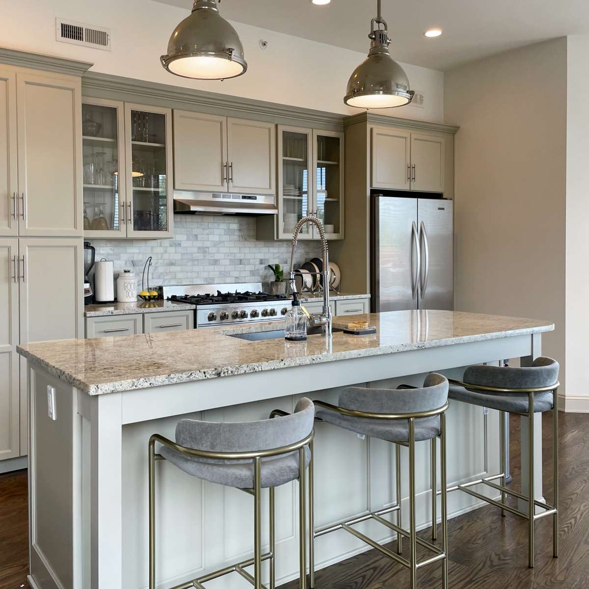 A modern kitchen features a large island with bar stools, grey cabinets, stainless steel appliances, and a white marble countertop.