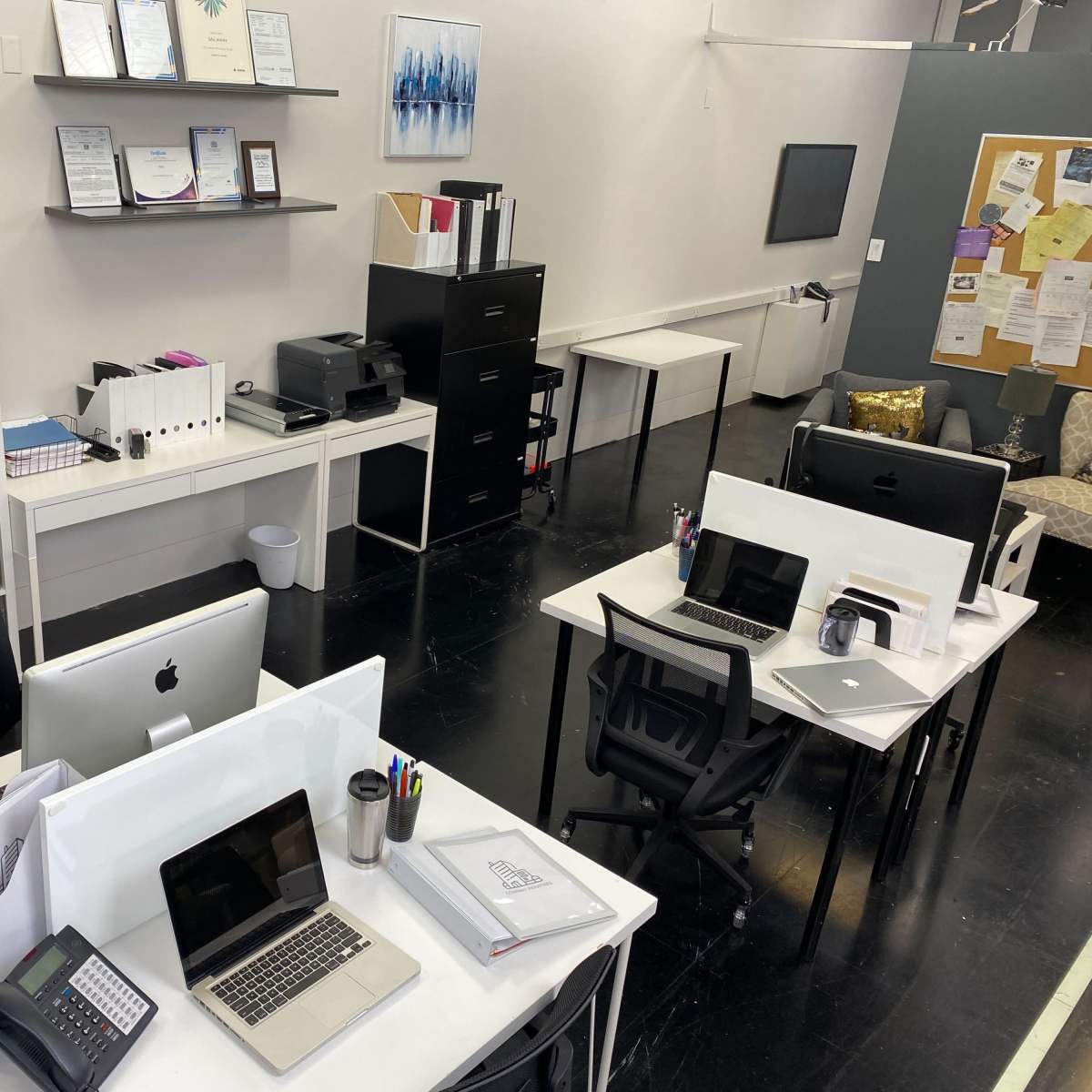 A modern office space with multiple workstations, a printer, filing cabinets, and a bulletin board.