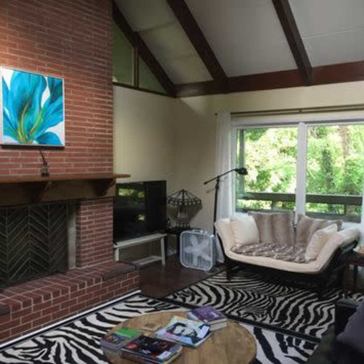 A living room with a brick fireplace, a large window overlooking greenery, a patterned rug, and a sectional sofa.