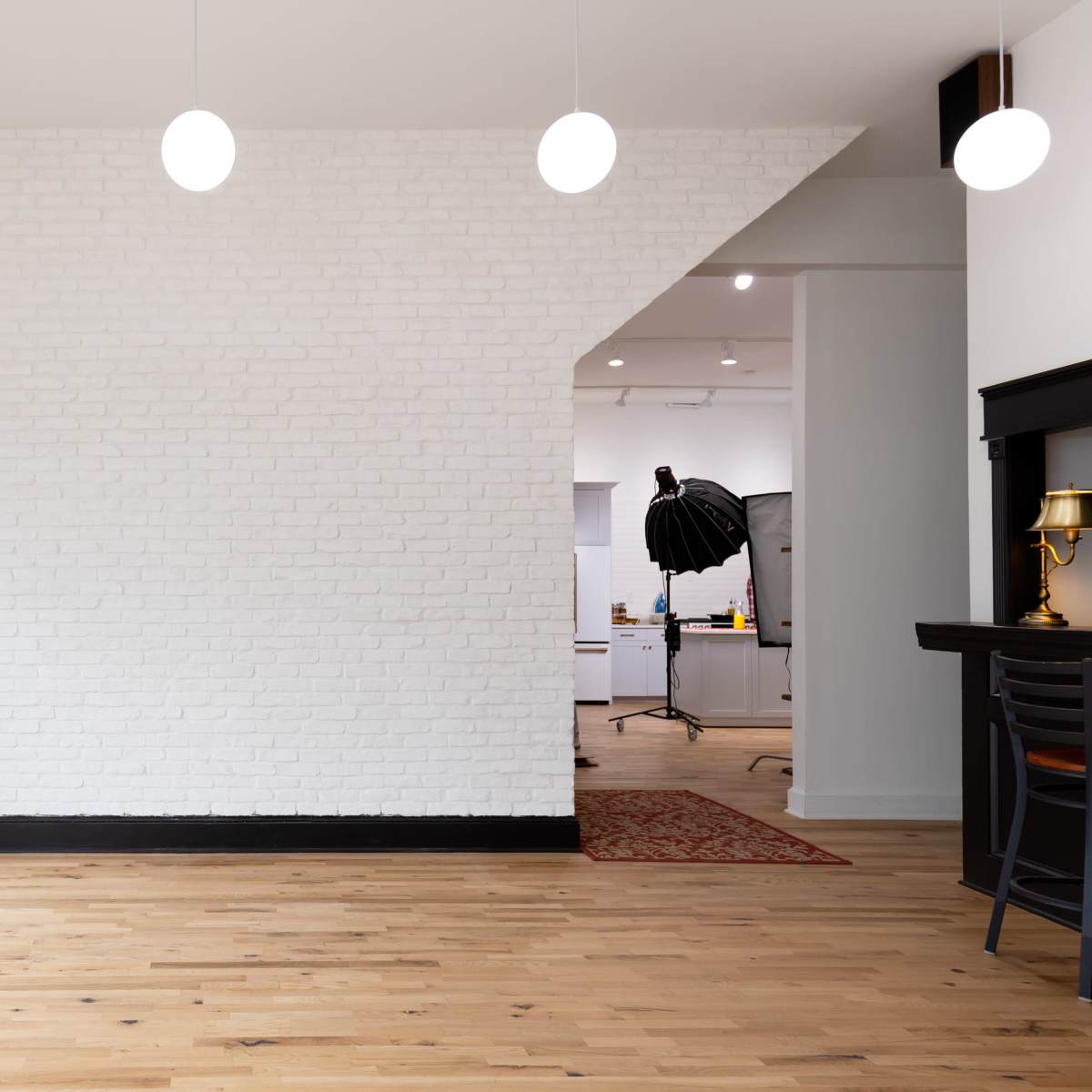 A spacious, well-lit room features a white brick wall, wooden floors, and hanging pendant lights, with a plant in a pot and a glimpse of office equipment in the background.