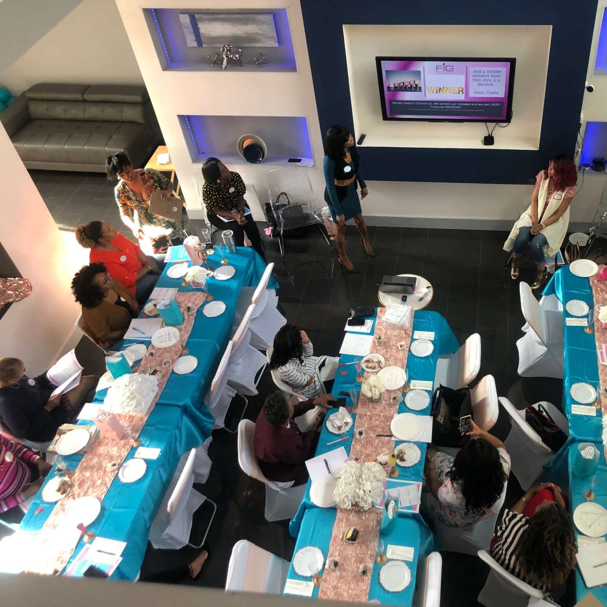 A group of people is seated at a long table covered with turquoise tablecloths in a modern room, with a presentation screen displaying information at one end.