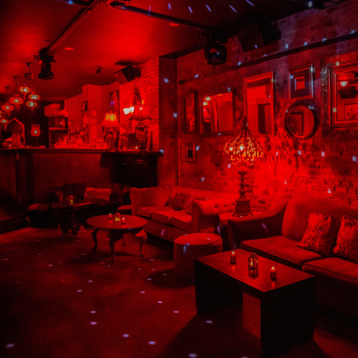 A dimly lit lounge area with red lighting, featuring sofas, a coffee table, and multiple mirrors on the walls.