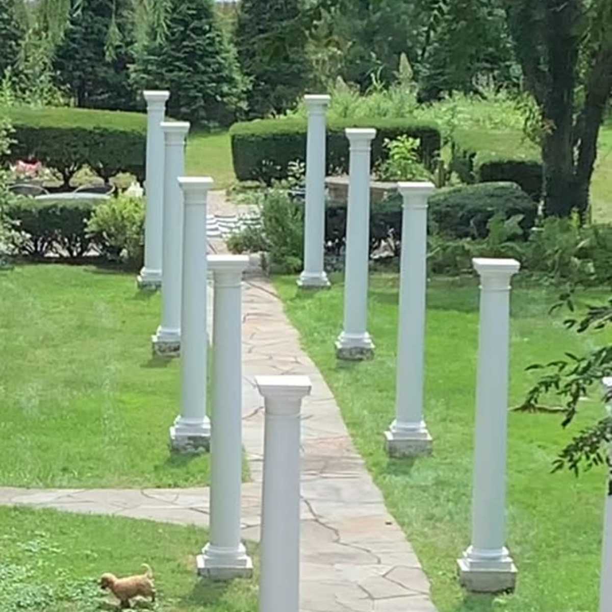 A paved pathway lined with white columns runs through a grassy area with neatly trimmed bushes and trees.