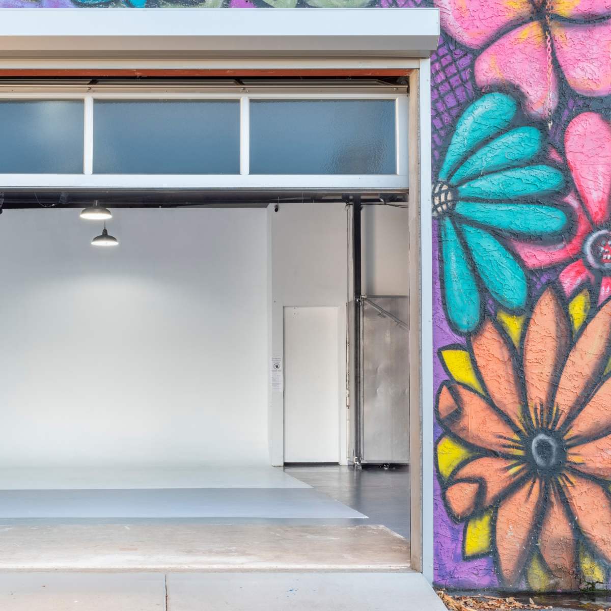 A large open garage door reveals a minimal interior space with plain walls and a polished floor, framed by vibrant graffiti on the outside.
