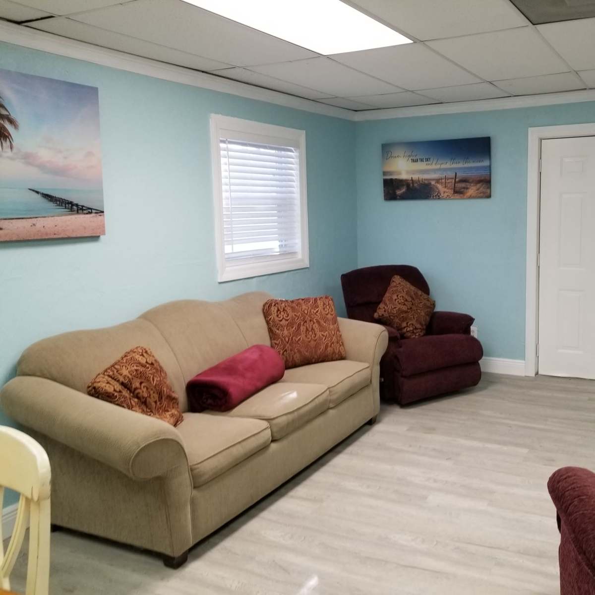 The room features a light blue wall with two armchairs, a sofa with decorative pillows, and framed beach landscapes on the walls.