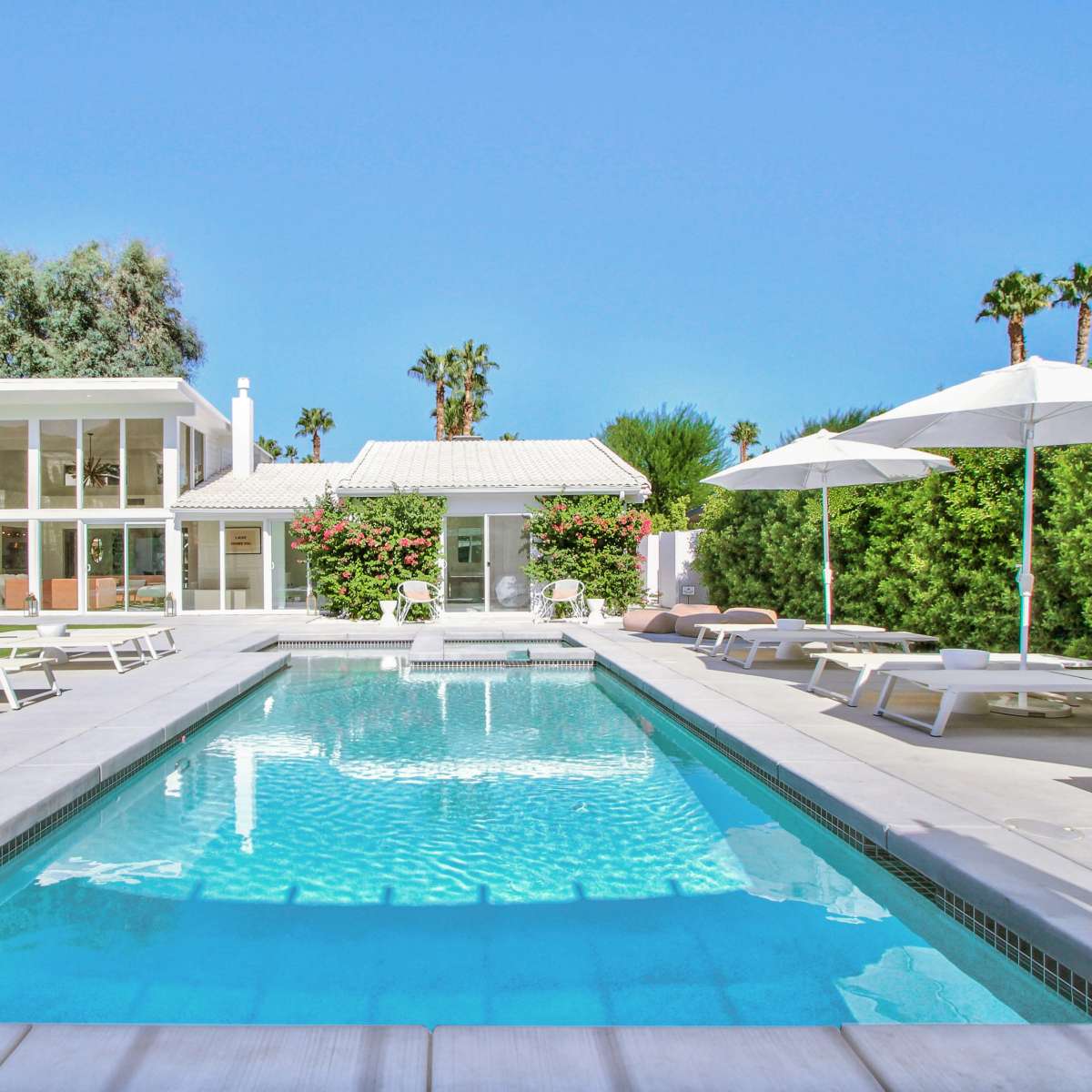 A modern pool area surrounded by lounge chairs, with a clear blue pool and a white house in the background, complemented by palm trees and landscaped greenery.