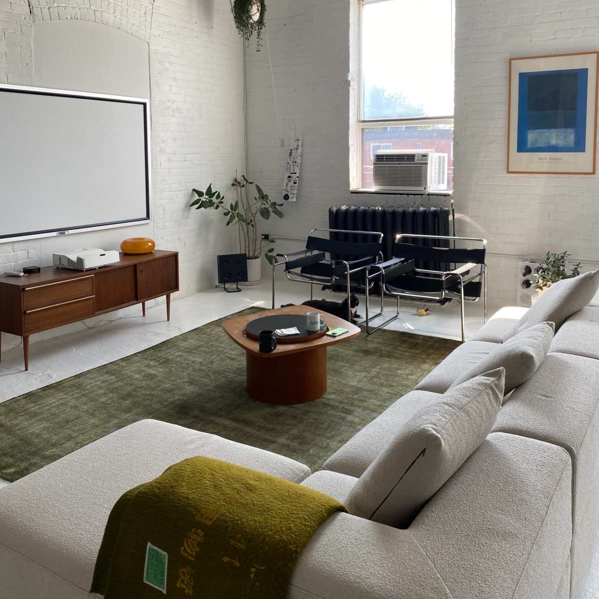 A bright, minimalist living room featuring a white sectional sofa, a wooden coffee table, and a wall-mounted projection screen.