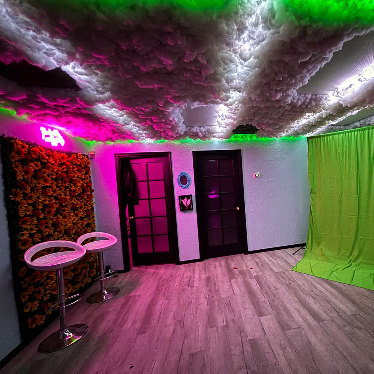 A room with a textured ceiling illuminated by colorful lighting, a green backdrop, two bar stools, and dual black doors.