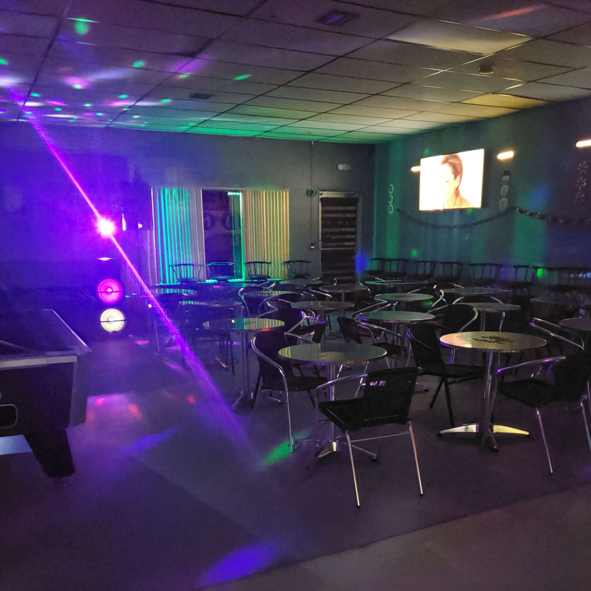 An empty lounge with tables and chairs illuminated by colorful lights and a screen displaying a video in the background.