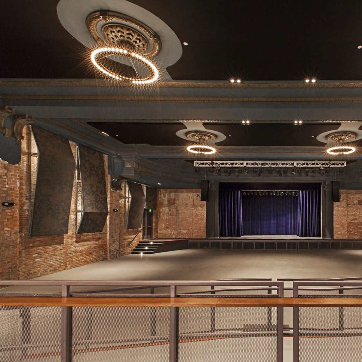 A spacious auditorium with exposed brick walls, a raised stage draped in blue curtains, and decorative lighting fixtures on the ceiling.