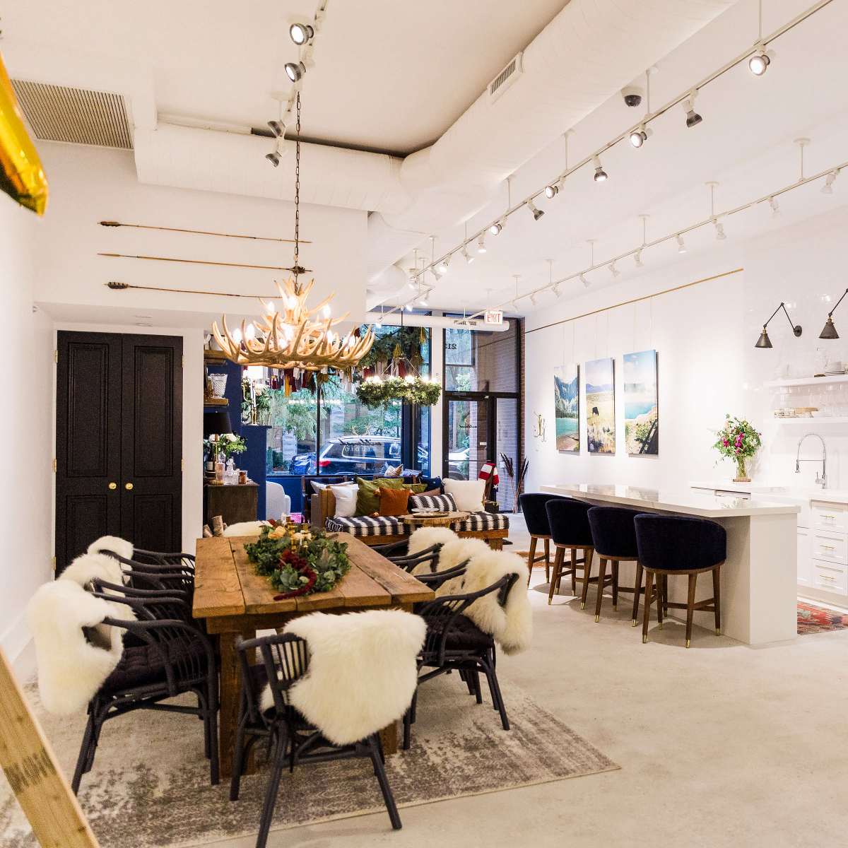 A modern interior of a café or restaurant featuring a long wooden dining table with upholstered chairs, a bar area with stools, and large windows allowing natural light.
