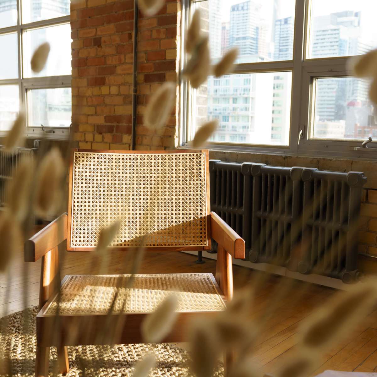 A wooden chair with a woven backrest is positioned in a bright room filled with large windows overlooking a cityscape.