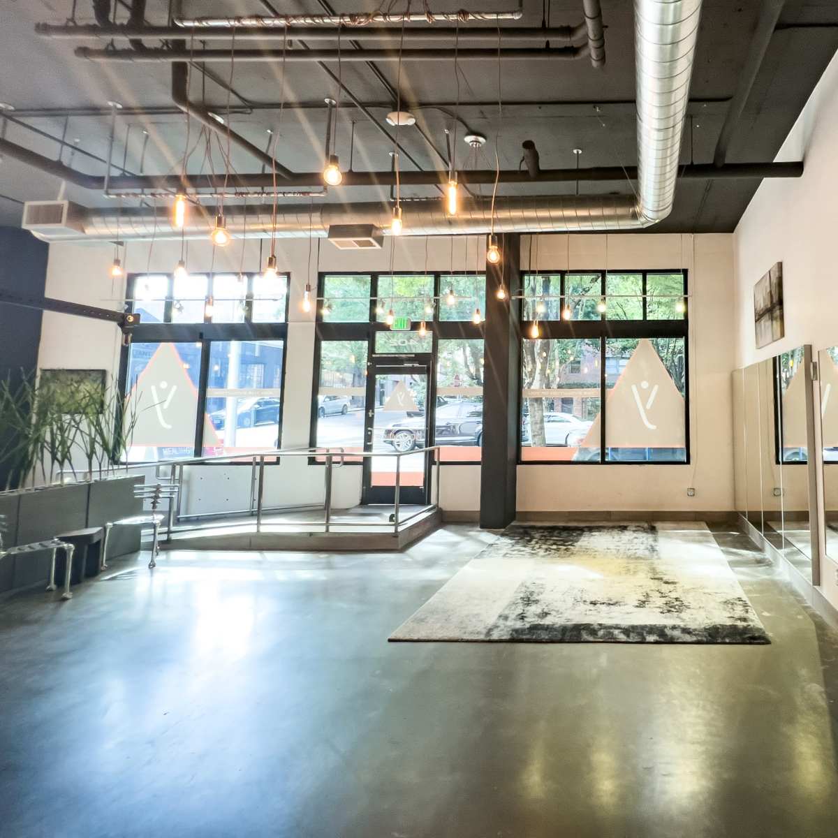 A spacious interior with a large mirror on one wall, industrial light fixtures, and large windows providing natural light.