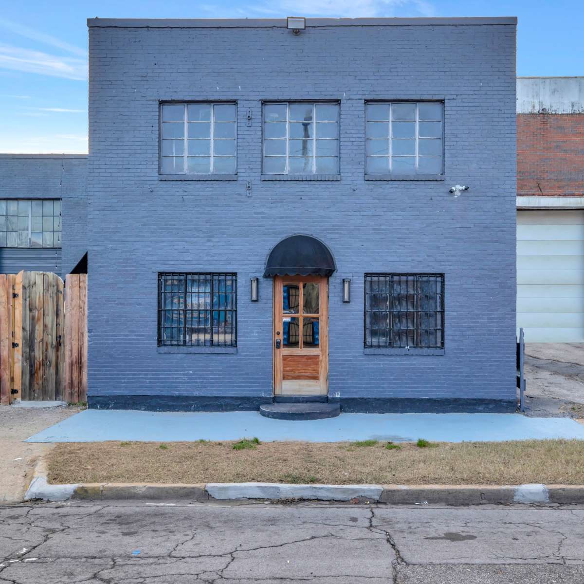 A two-story gray building with a wooden door and large front windows, situated next to a wooden fence and a brick warehouse.
