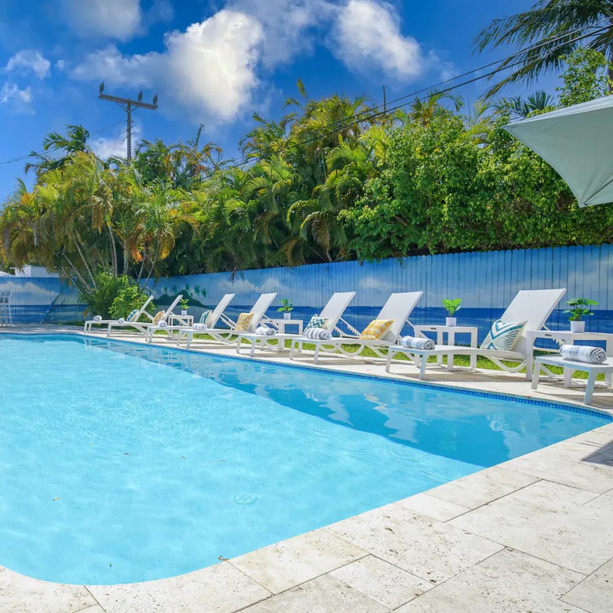 A clear swimming pool is surrounded by lounge chairs and palm trees under a blue sky.