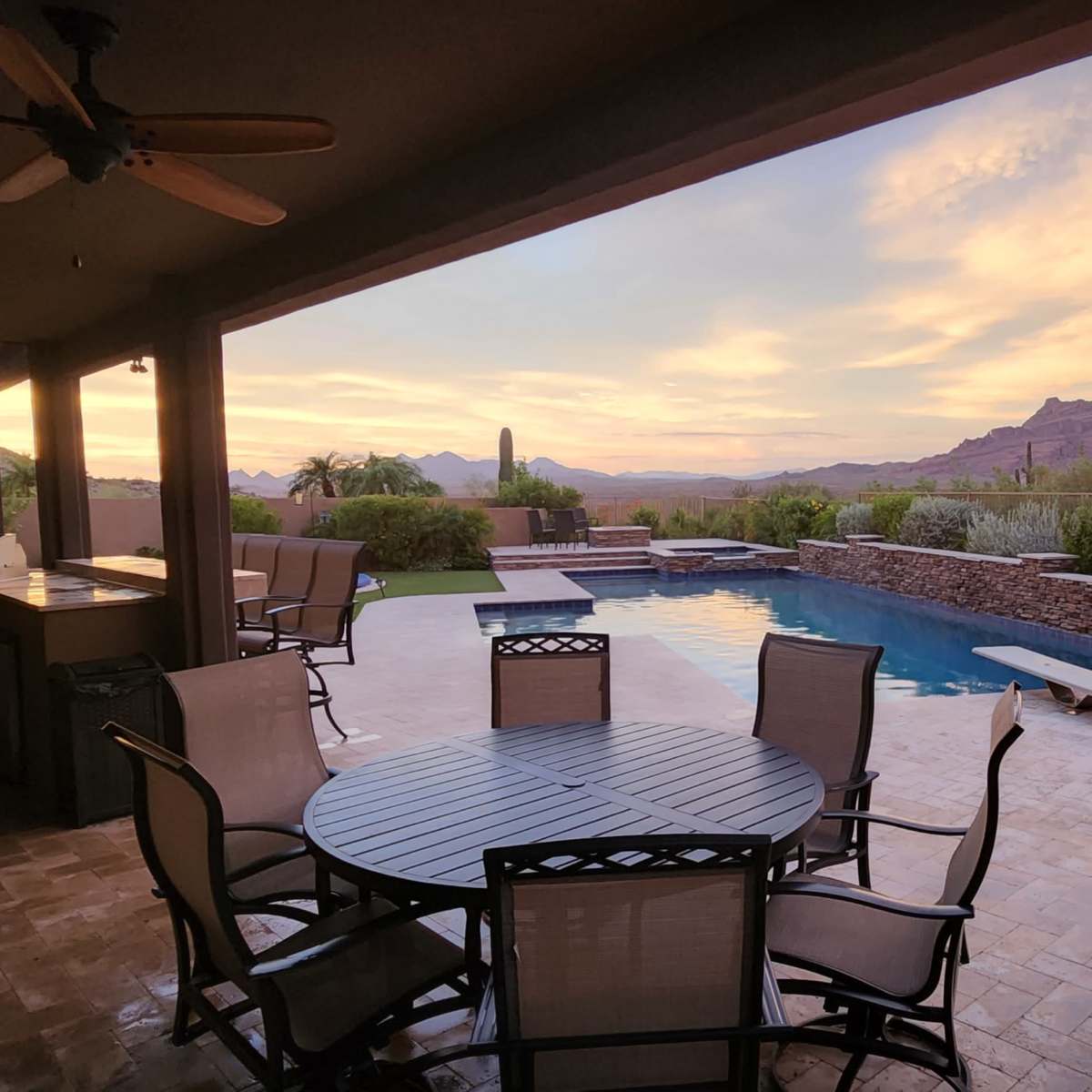 A covered patio with a dining table and chairs overlooking a swimming pool and mountains at sunset.