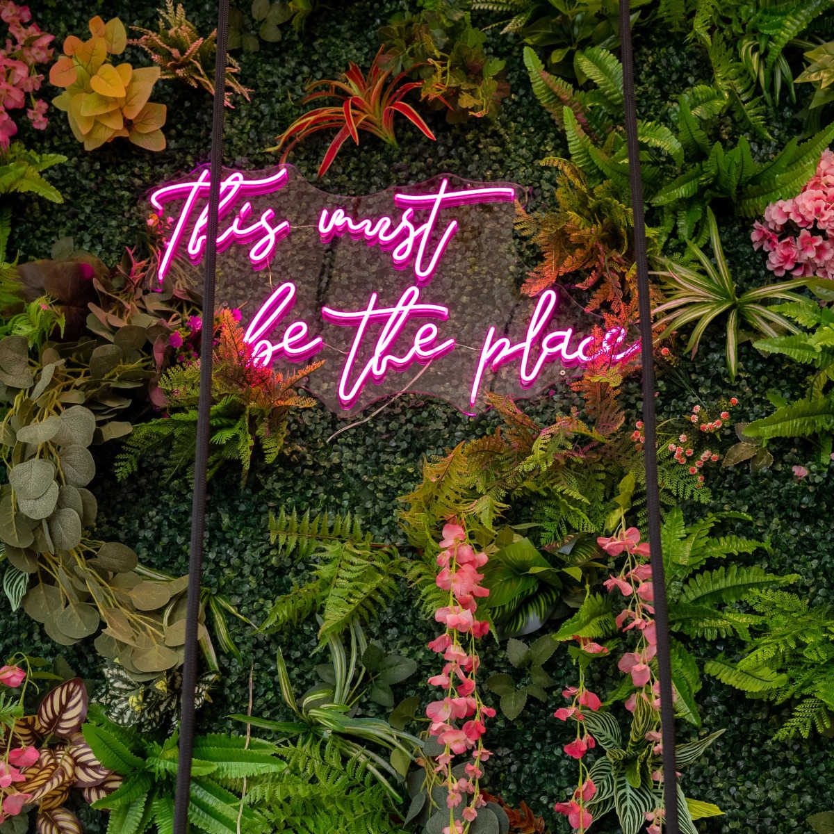 A swing hangs in front of a vibrant green wall adorned with various plants and a neon sign that reads, "It must be the place."