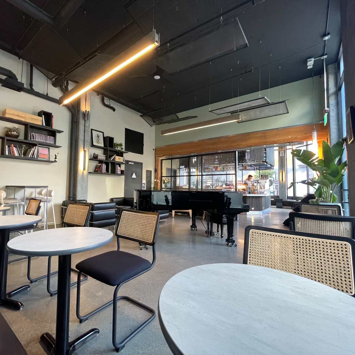 The interior of a modern café features tables and chairs, a piano, and large windows that provide a view of the outside.