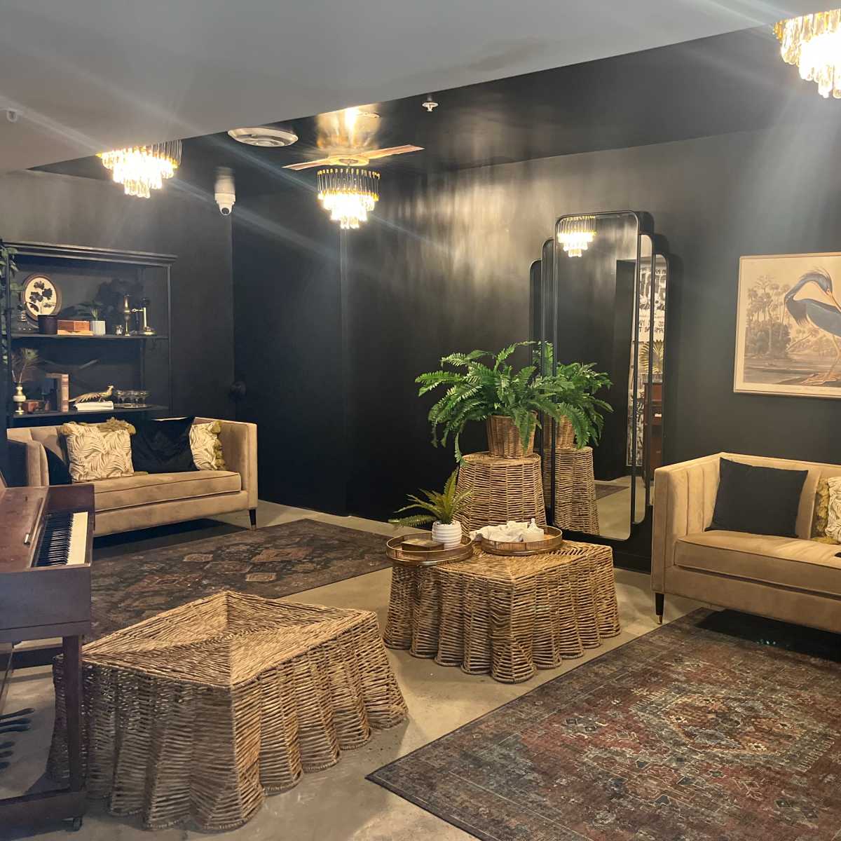The room features two beige sofas, a piano, a large mirror, and several rattan furniture pieces, all set against dark walls and illuminated by chandelier lighting.