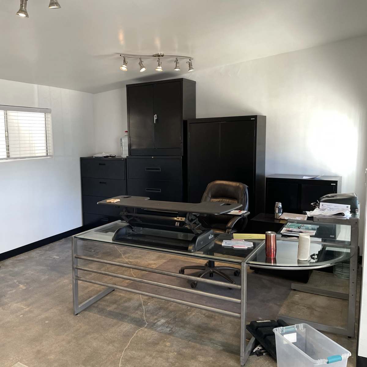 A minimalist office space featuring a glass-top desk, a black office chair, filing cabinets, and a mini fridge, with ample natural light coming through a window.