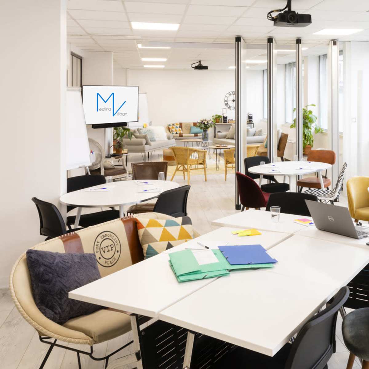 A modern, open-plan workspace featuring several tables and chairs, a projector, and windows allowing natural light.