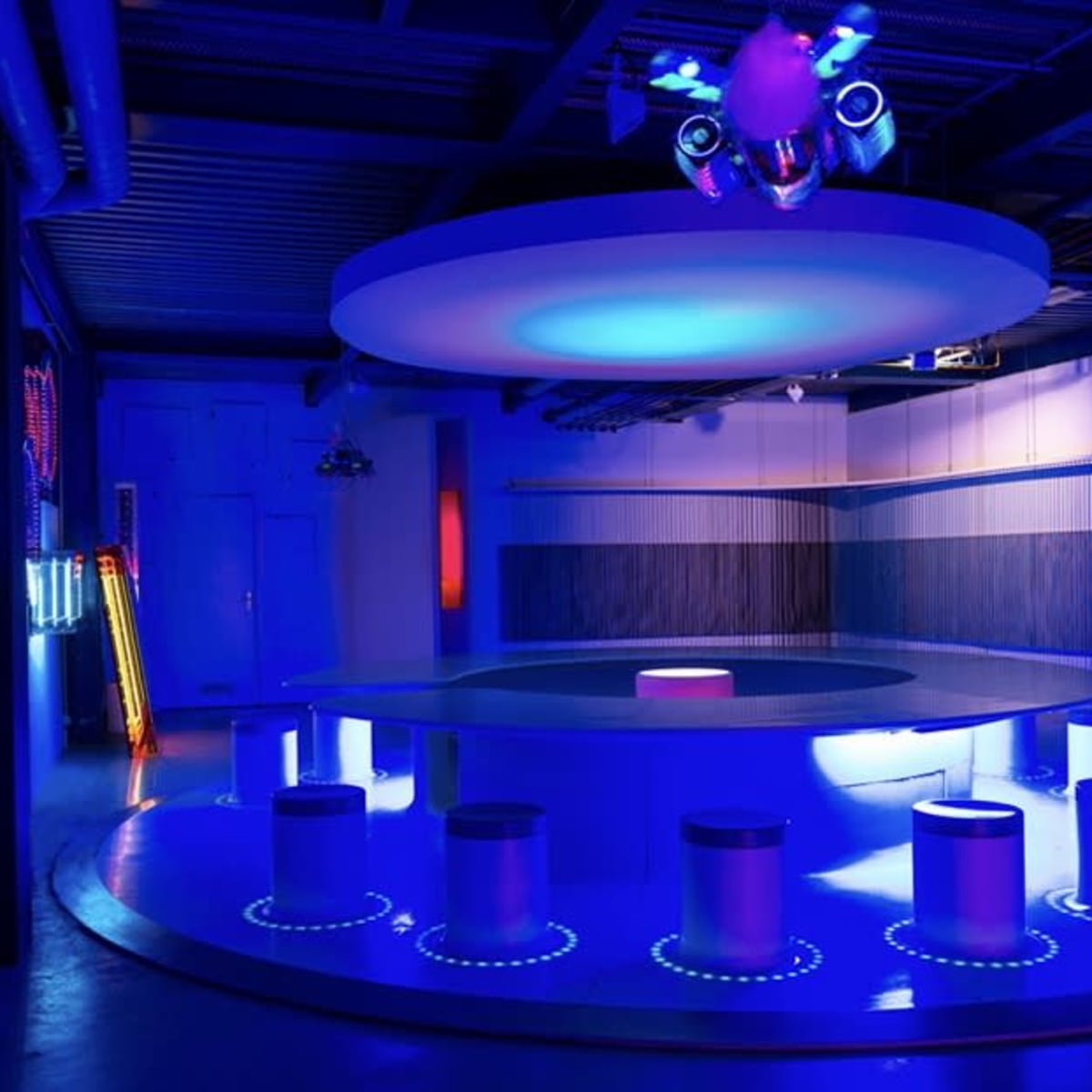 A modern, dimly lit lounge with circular seating and illuminated surfaces, featuring blue lighting and abstract wall art.