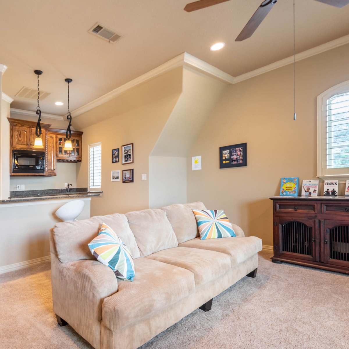 A cozy living room features a light beige sofa with colorful pillows, a small kitchenette, and a wooden cabinet, all set against a softly painted wall with a window.