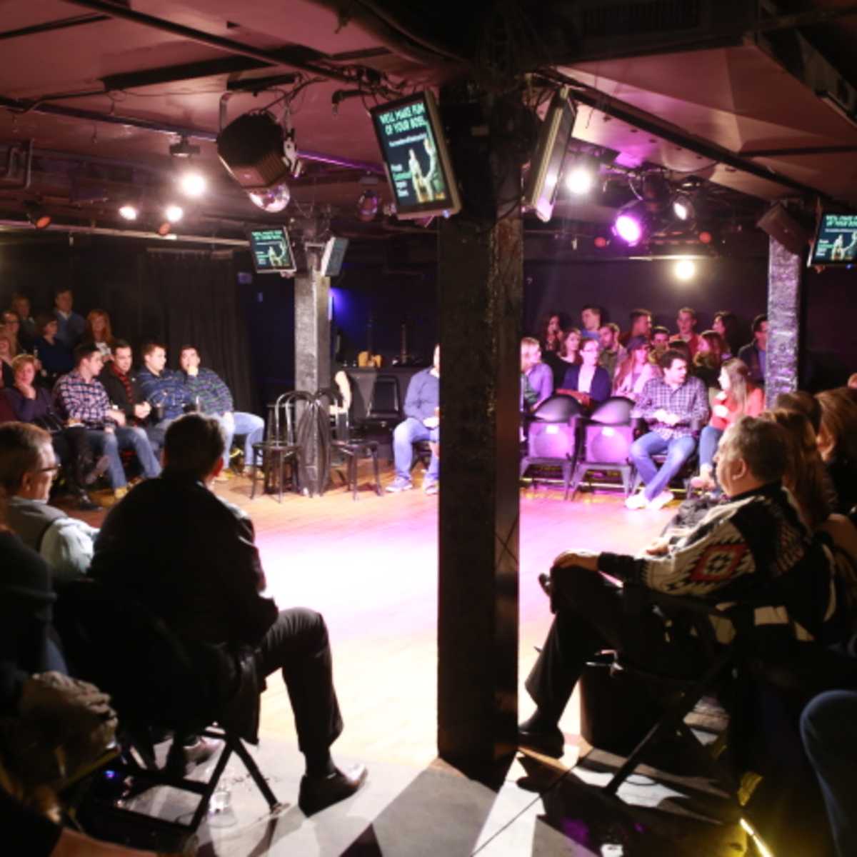 A comedy club audience is seated in rows facing a small stage with multiple screens displaying information.