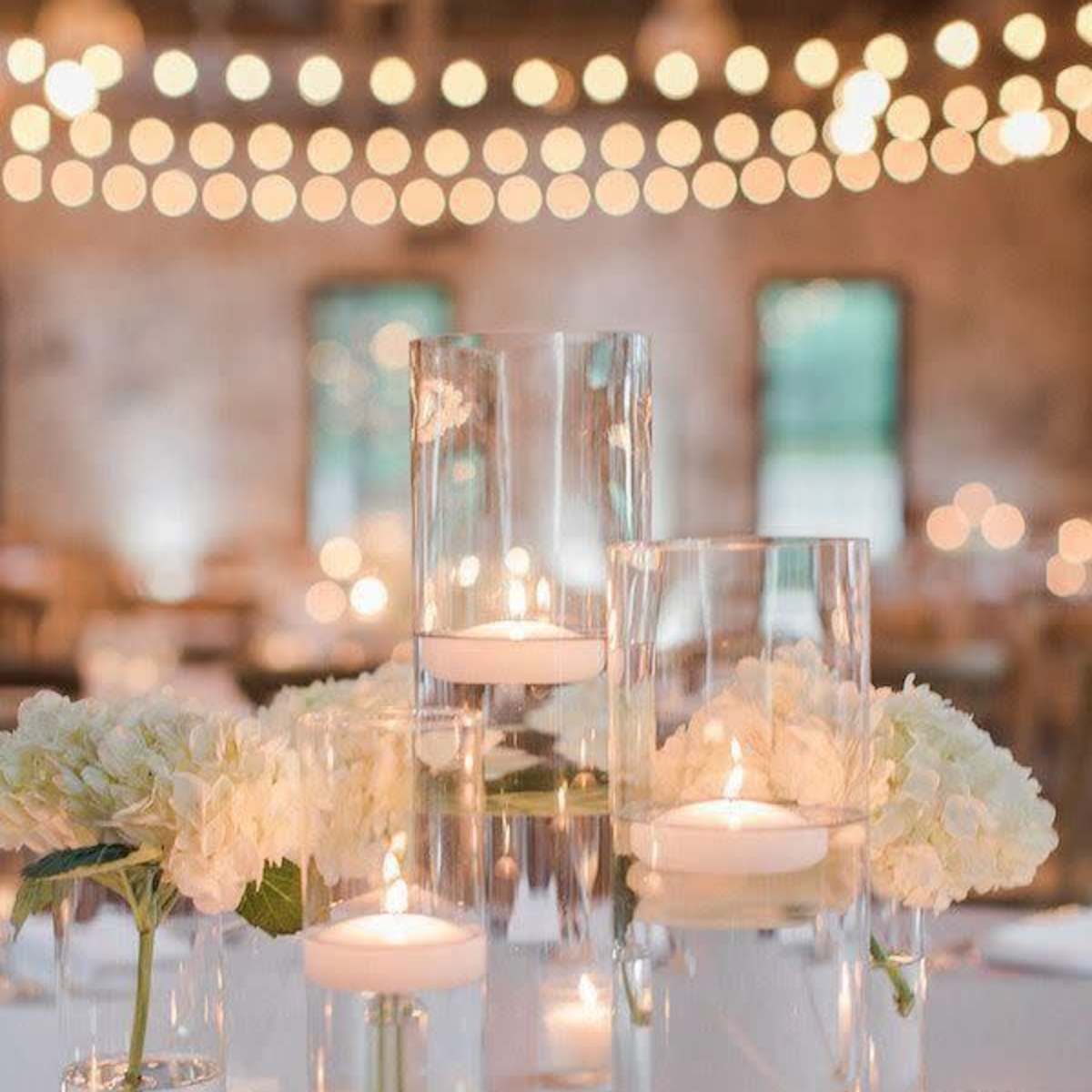 A table setting adorned with glass vases of varying heights, each containing floating candles and surrounded by white hydrangeas, all illuminated by soft, warm string lights in the background.