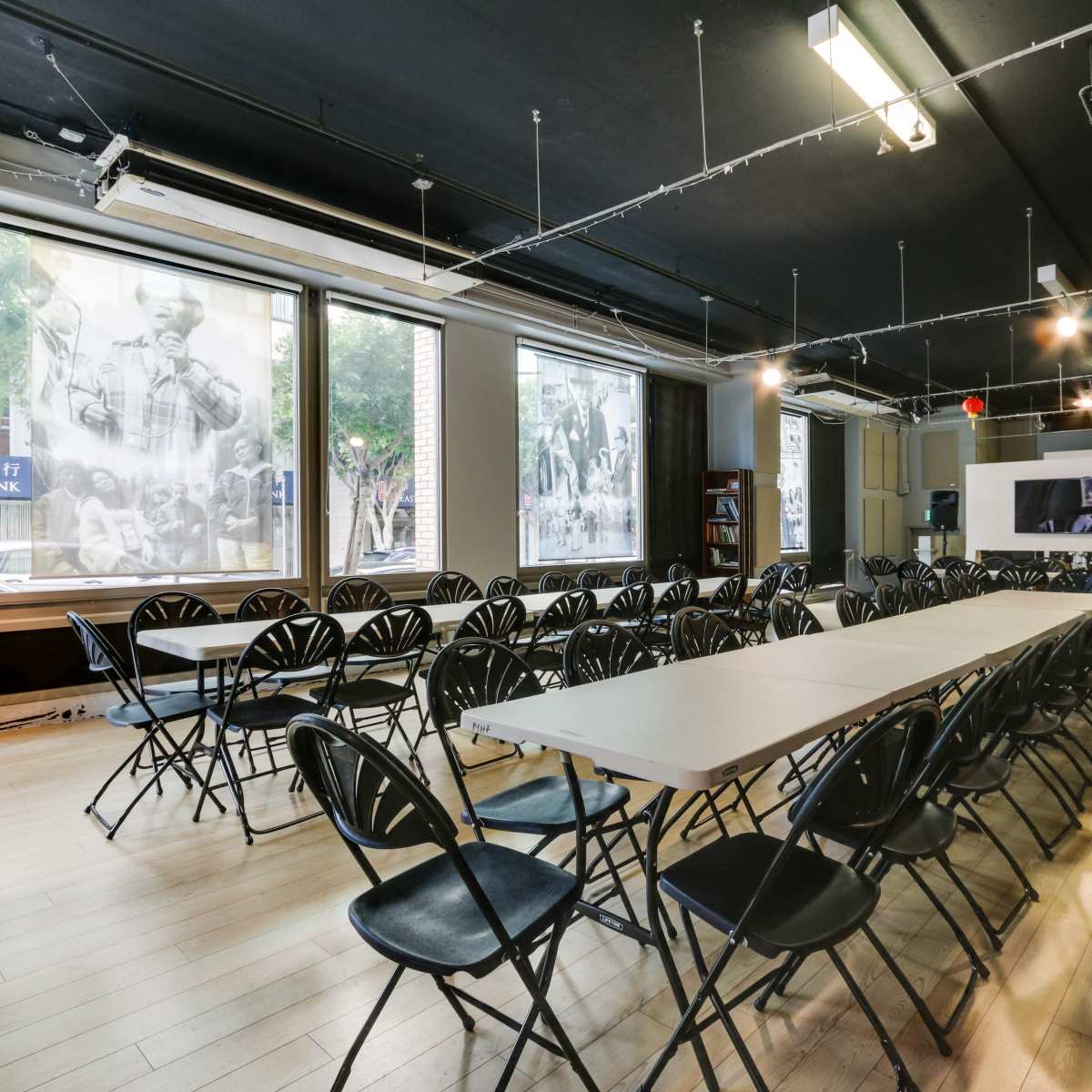 A spacious room is set up with rows of folding chairs facing a long table, surrounded by large windows showcasing black-and-white photographs.