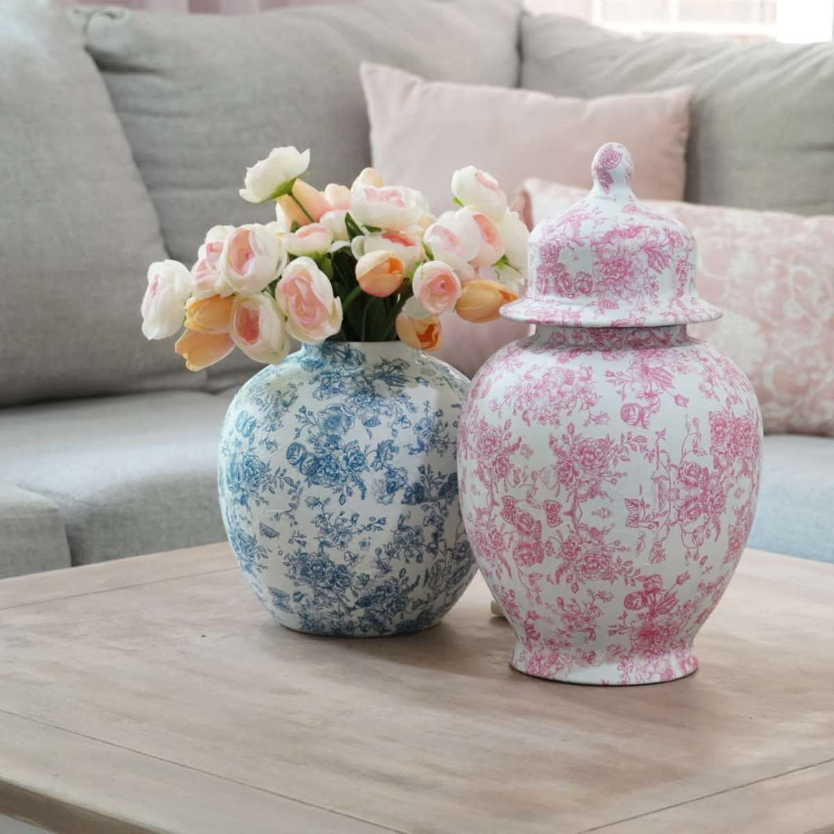 Two decorative vases, one blue and white and the other pink and white, are placed on a wooden coffee table beside a floral arrangement in a living room with a light-colored sofa.