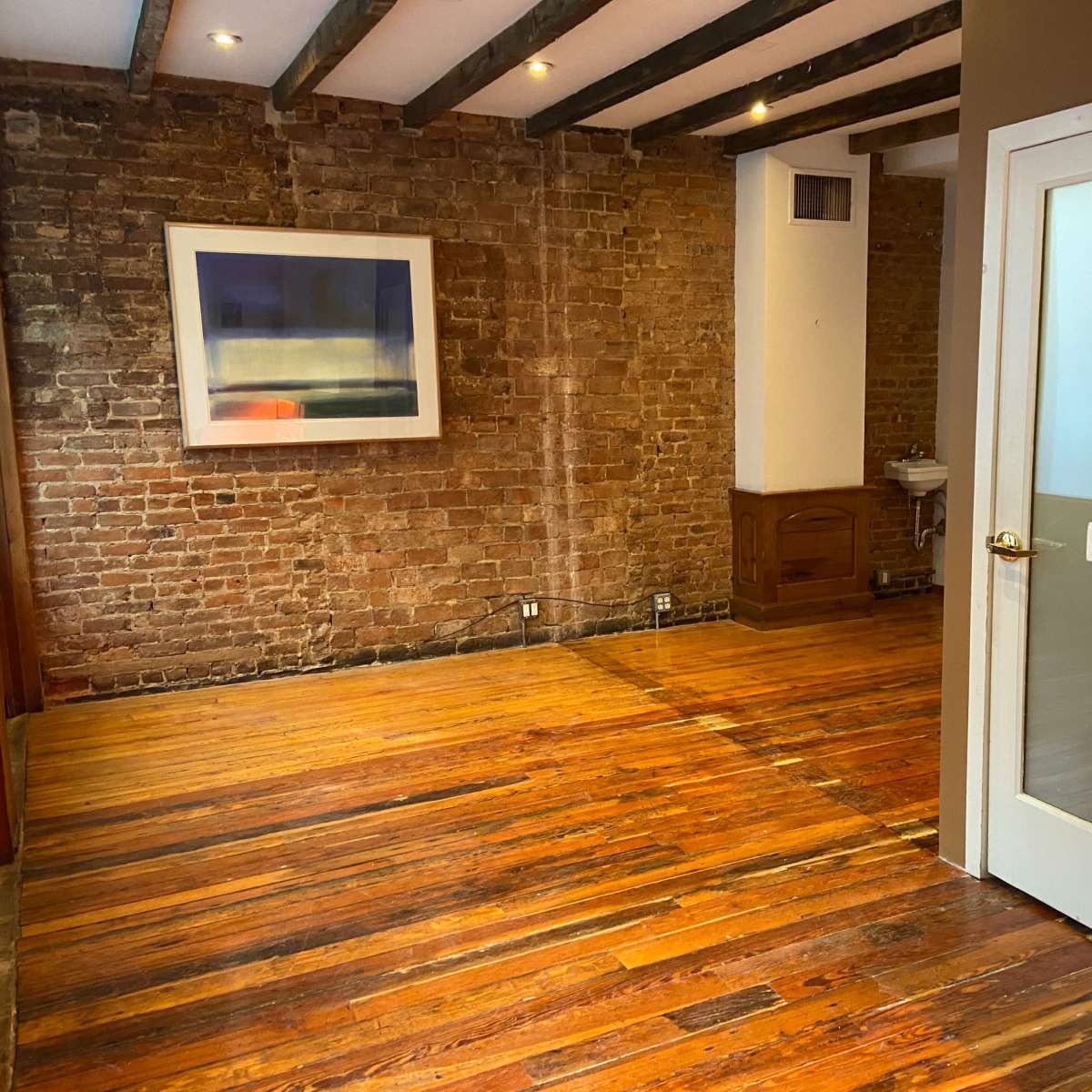 A spacious room with exposed brick walls, hardwood floors, and a doorway leading to another area.