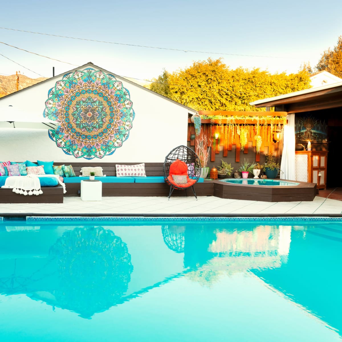 A backyard pool area features a colorful mural on the wall, surrounded by comfortable seating and a red accent chair near the water's edge.