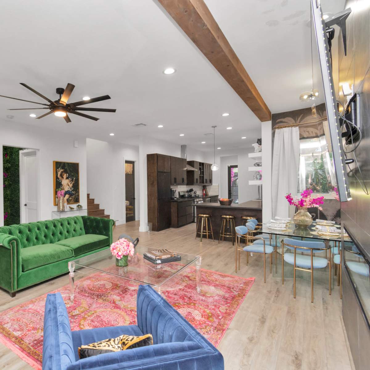 A spacious living room features a green velvet sofa, a glass coffee table, and a modern kitchen with dark cabinetry in the background.