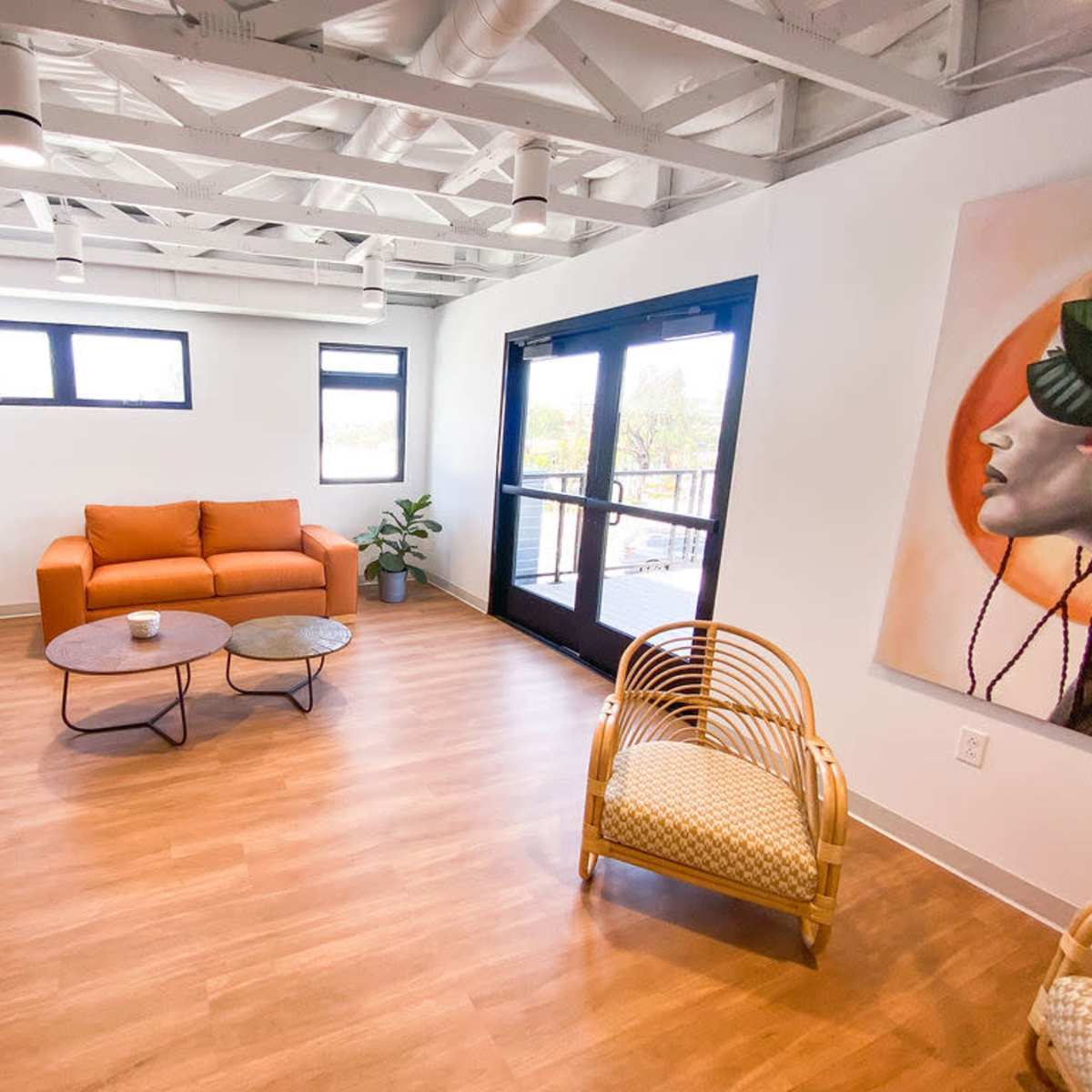 A modern interior space features a brown couch, a round coffee table, and a wicker chair, with large windows and wooden flooring.