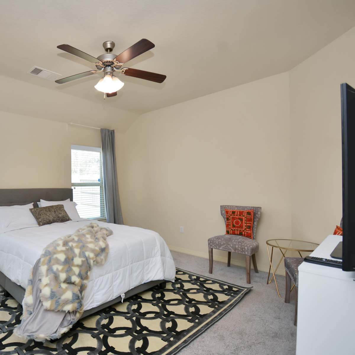 A bedroom with a queen-sized bed, a ceiling fan, two windows, a chair in the corner, and a decorative rug on the floor.