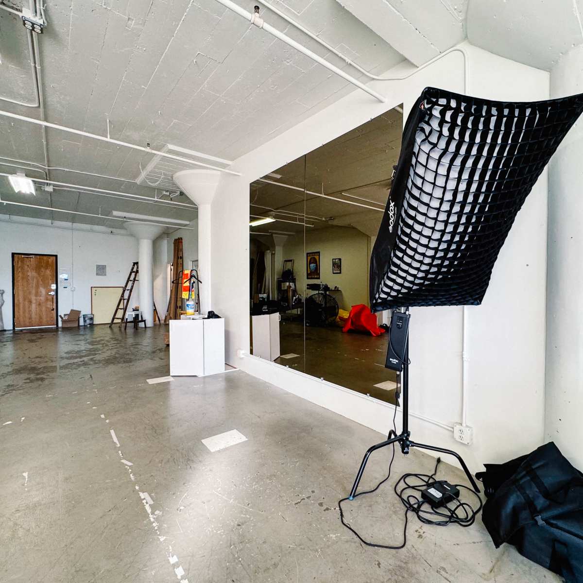 A spacious, unfinished studio with a large mirror, lighting equipment, and scattered furniture.